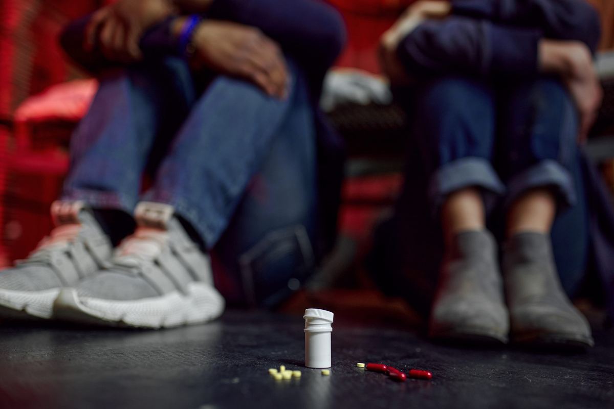A pill bottle and loose capsules on the floor in front of two people seated on a couch.
