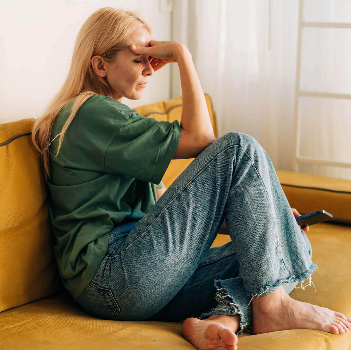 A distressed young woman patient of Balanced Mental Health of Arizona sitting on her couch resting her head in her hands.