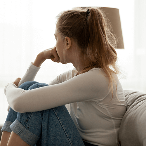 A young woman patient of Balanced Mental Health of Arizona sitting on a couch with her head resting on her chin looking out a window.