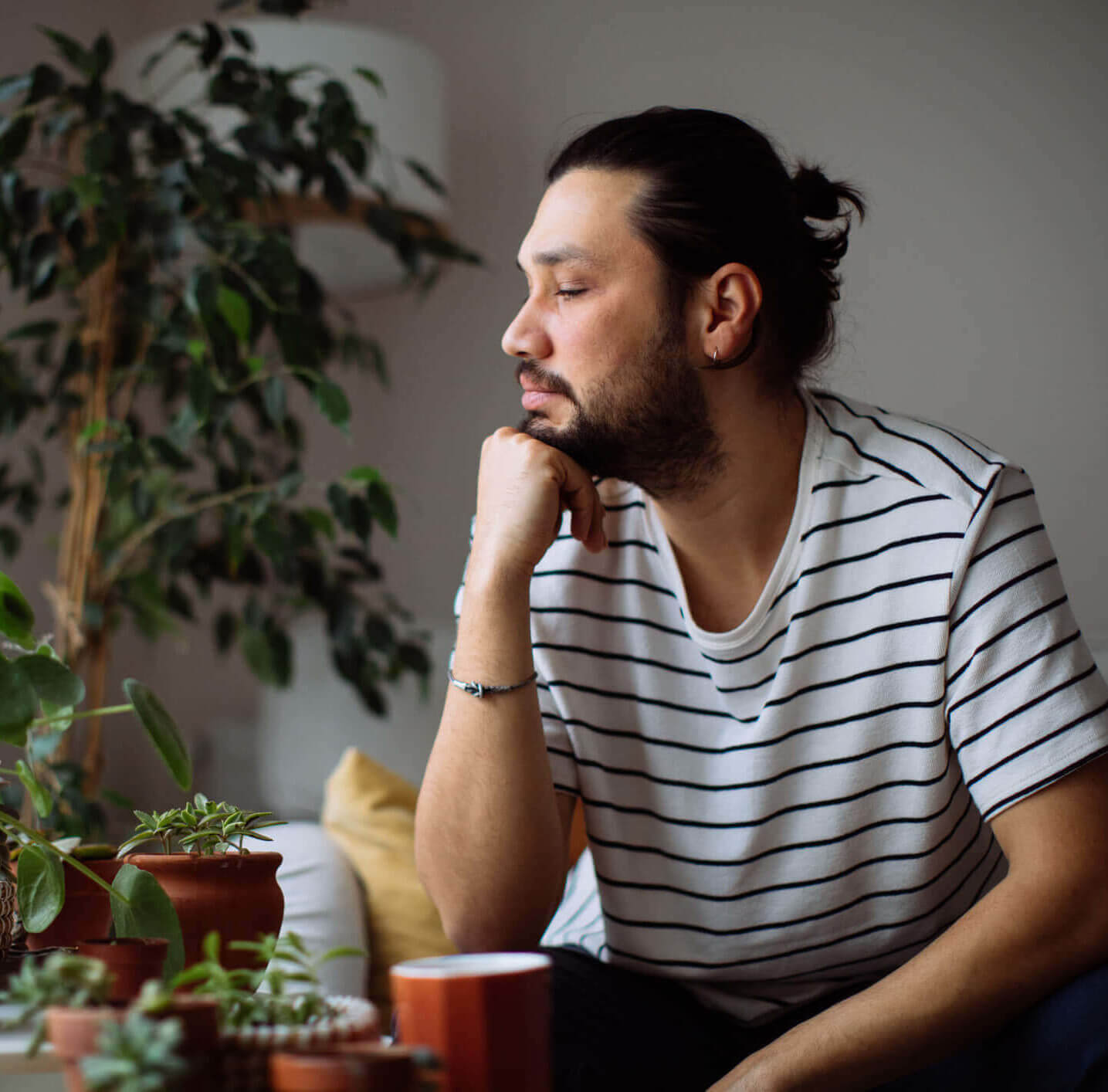 A young man, a patient of Balanced Mental Health of Arizona, wearing a striped T-shirt resting his head on his chin while holding a cup of coffee and looking serene.