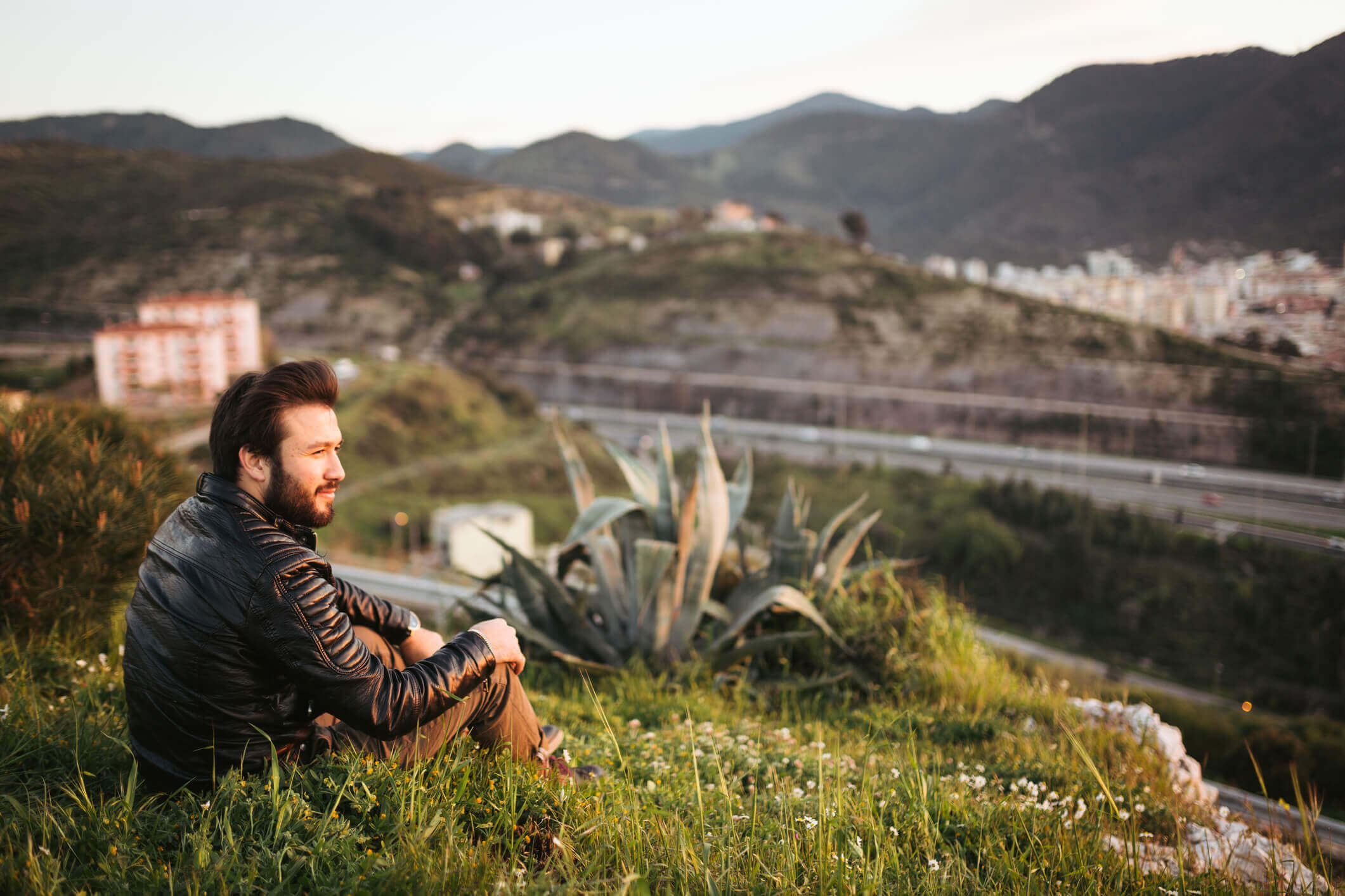 Man wearing a leather jacket sitting on a grassy hilltop.